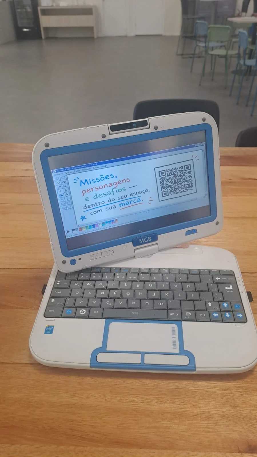Netbook branco e azul de 2012 sobre uma mesa, com a tela girada para frente exibindo no Paint uma apresentação da Guilda de Aventureiros e um QR code.