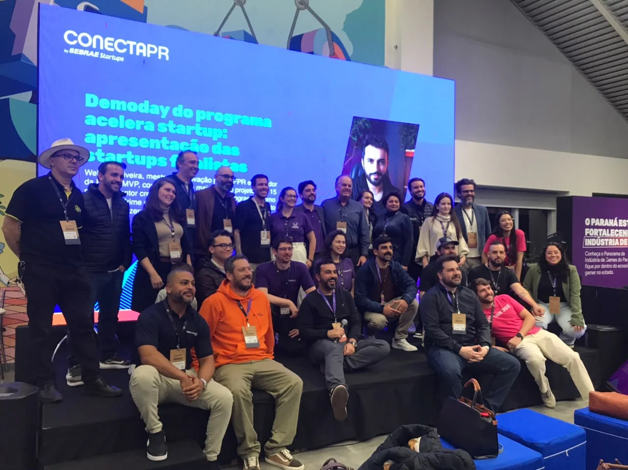 Grupo de participantes do Acelera Validação reunido diante do palco do Demoday do ConectaPR, com um telão azul ao fundo.
