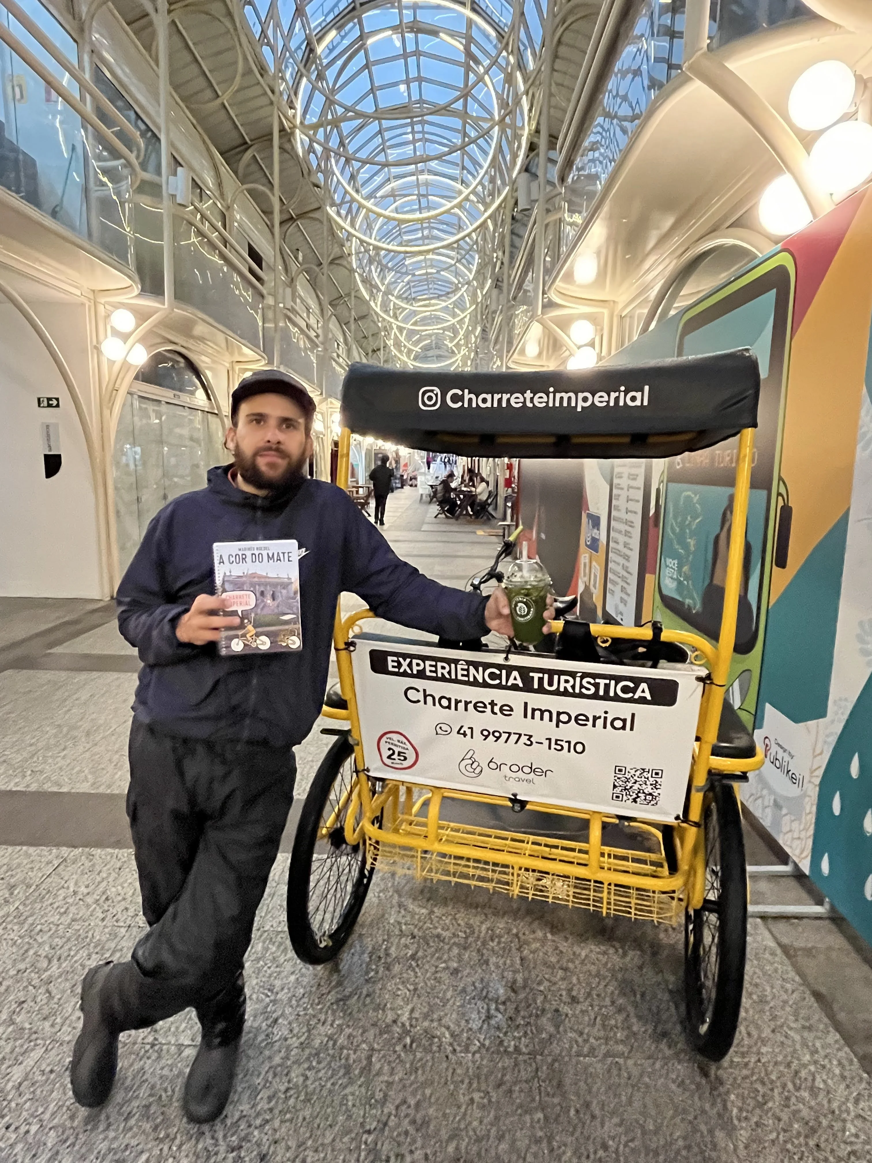 Leandro Roedel accanto a una bicicletta cargo gialla di Charrete Imperial, in una galleria di Curitiba, con la pubblicazione A Cor do Mate e una calebasse di chimarrão.